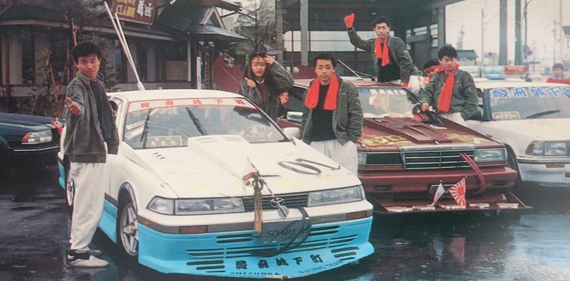 Group of racers with colorful custom cars and red scarves posing in a parking lot in Japan.