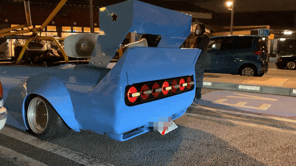 Modified blue car with a large rear wing and unique taillights in a parking lot at night, with Japanese city background.