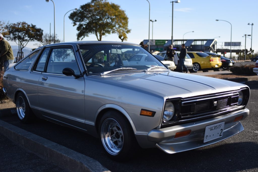 Classic silver car with Japanese license plate at a gathering on a sunny day. Trees and people in the background.