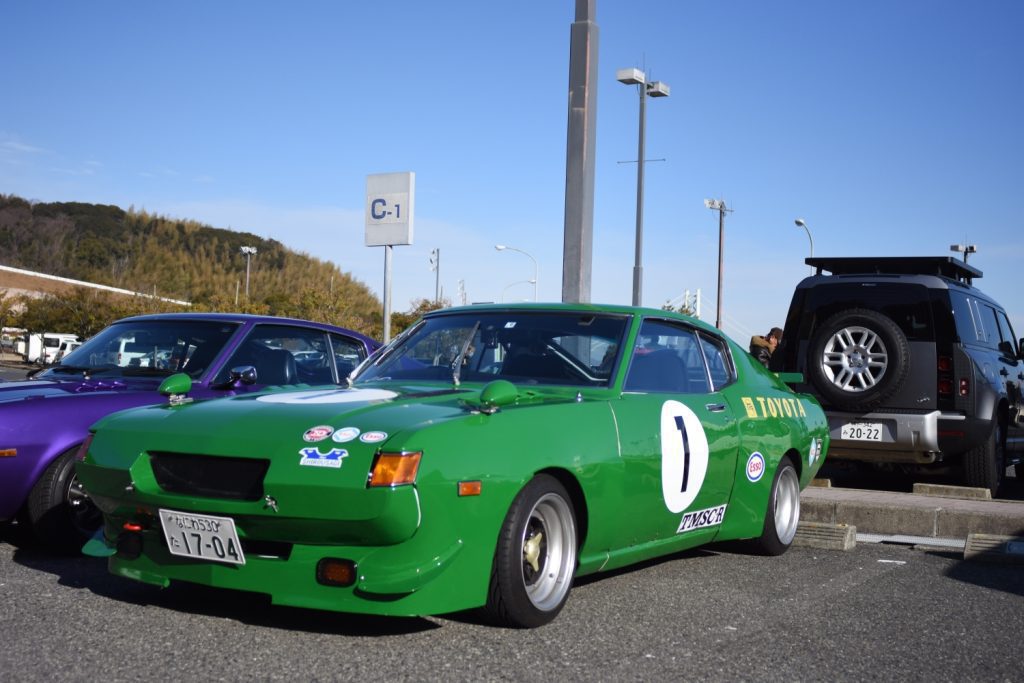 Vintage green Toyota race car in a parking lot with a license plate reading 17-04, parked near a purple car and an SUV.