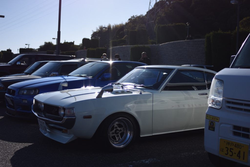 Classic Japanese cars parked, including a white vintage Toyota Celica, a blue Nissan Skyline, and a white utility van.