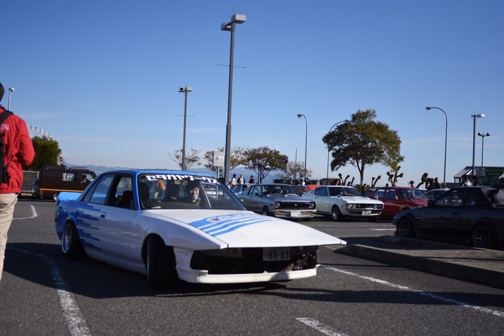 Group of vintage cars including a white modified car with a Japanese license plate at an outdoor car meet. #車集まり (Car gathering).