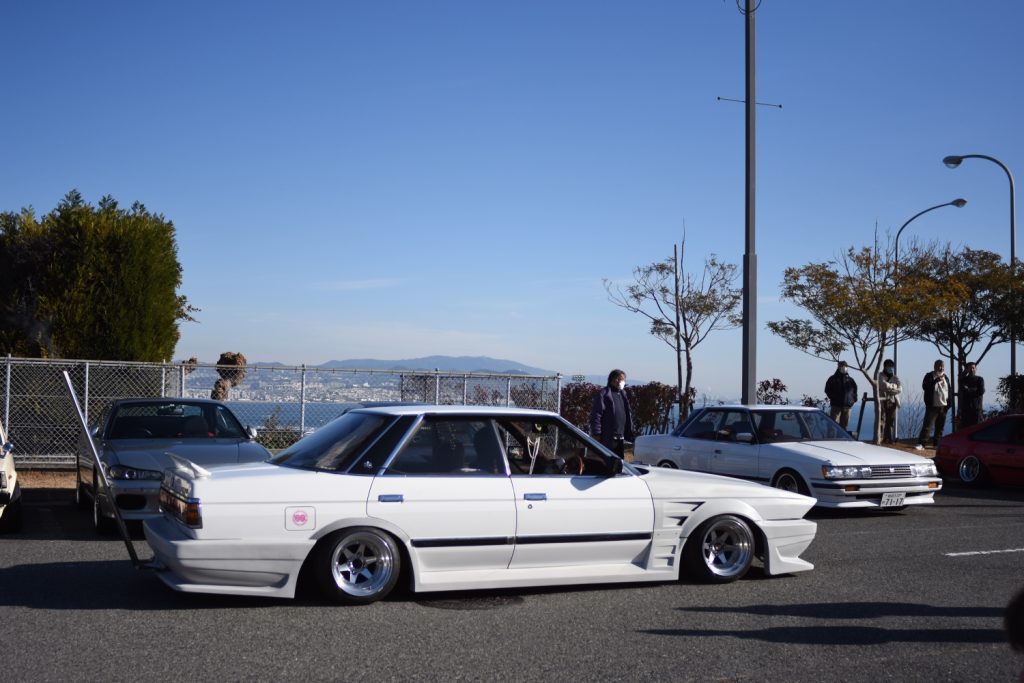 Modified white Japanese car at a car meet, with Mount Fuji in the background, sunny day, people observing parked cars.