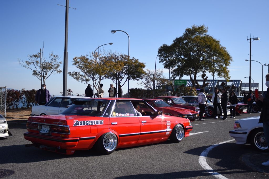 Red modified car with Bridgestone logo at an outdoor car meet in Japan, surrounded by other cars and people.