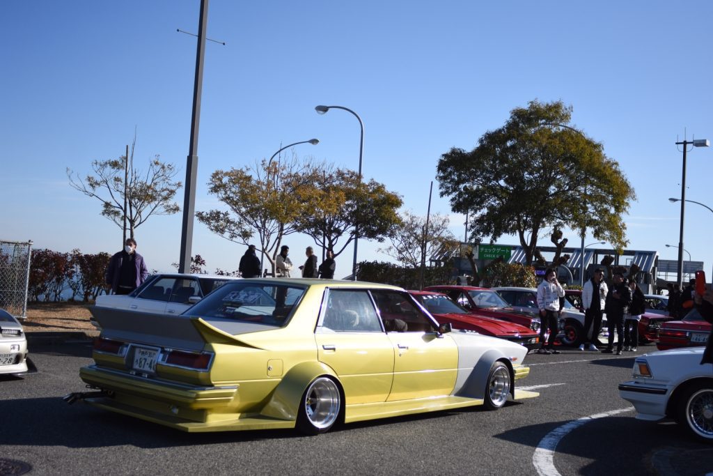 Yellow modified car at a Japanese car meet, with people and other cars in the background on a sunny day.