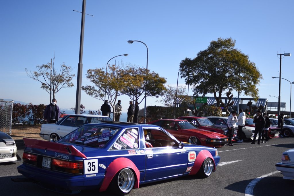 Japanese custom cars gathered at a car meet with onlookers under a clear blue sky.