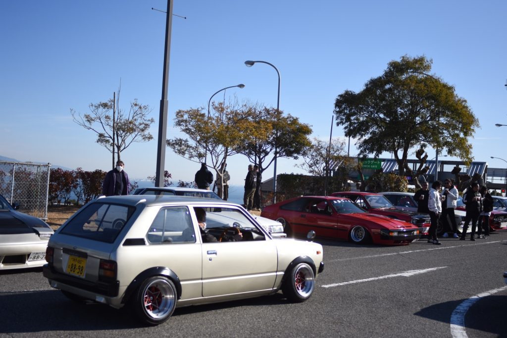 Vintage Japanese cars at a car meet, featuring a beige hatchback and red sports car, with people and trees in the background.