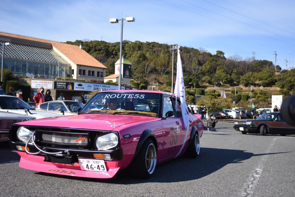 Bright pink classic car with Route55 banner in a Japanese car meet event, featuring text ロードフィフティーフィフ on the front bumper.