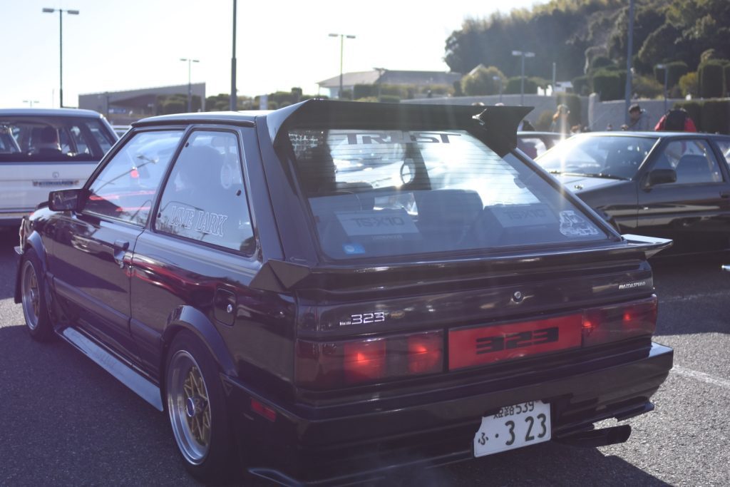 Modified classic Mazda 323 hatchback with Lone Lark decal and custom wing at a car meet in Japan. License plate: 3 23.