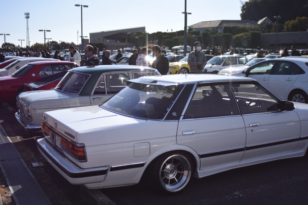 Car enthusiasts gather at a car show displaying classic and modern vehicles under clear skies in a parking lot.