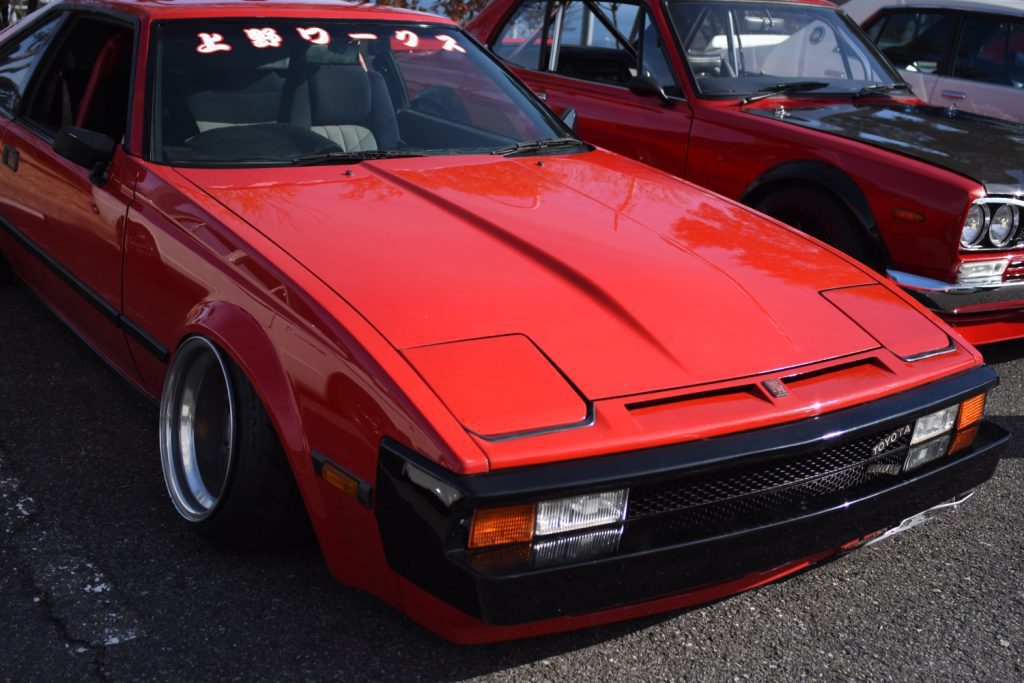 Bright red classic Toyota sports car with Japanese decal at a car show. Japanese text translation: Work