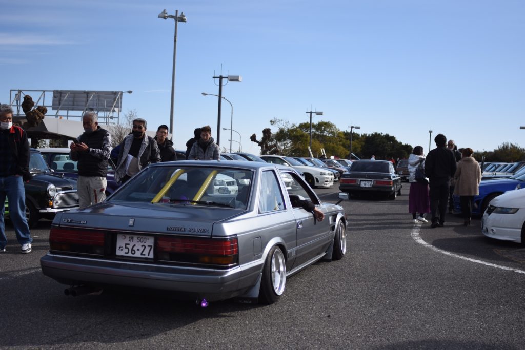 A classic Toyota Soarer with modifications at a car meet in Japan, surrounded by car enthusiasts and other vehicles.