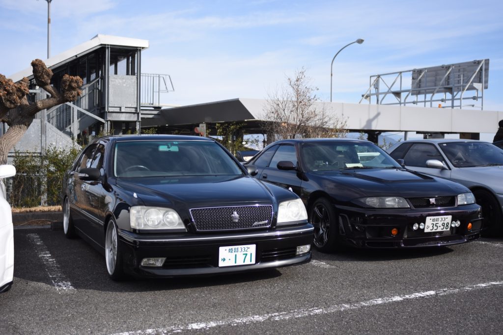 Two classic black Japanese sports cars parked side by side in a parking lot on a sunny day.