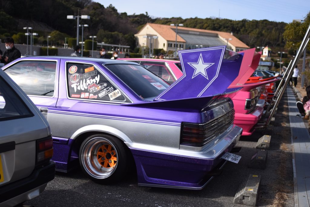 A lineup of customized Japanese cars featuring large rear wings and colorful paint jobs at a car event.