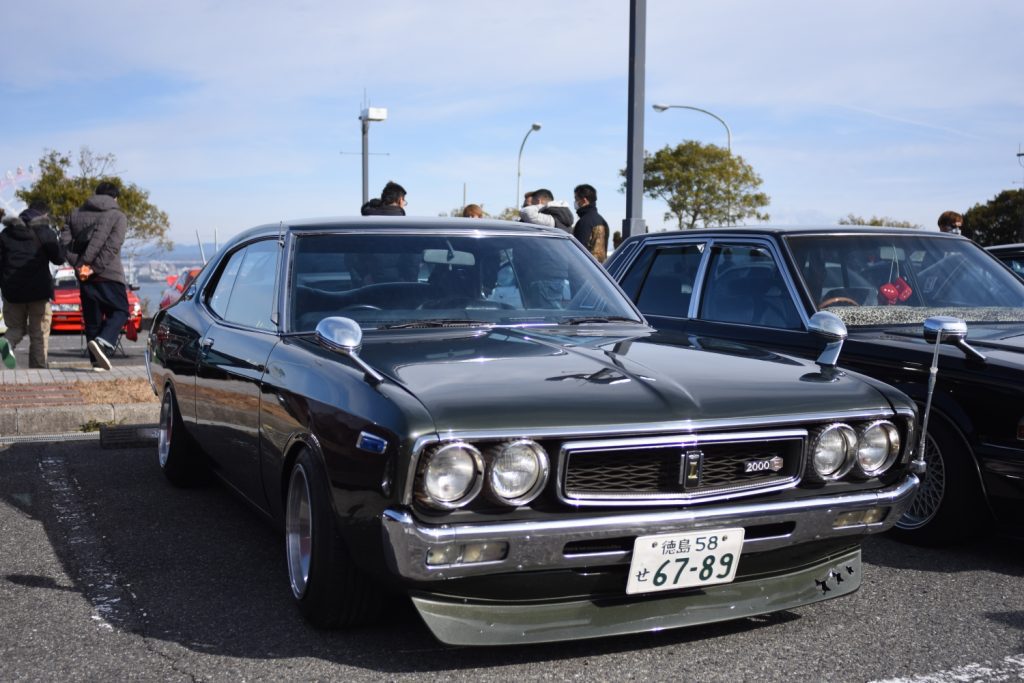 Classic Japanese car at auto meetup with green license plate from Tokushima, Japan, number 67-89, with people in the background.