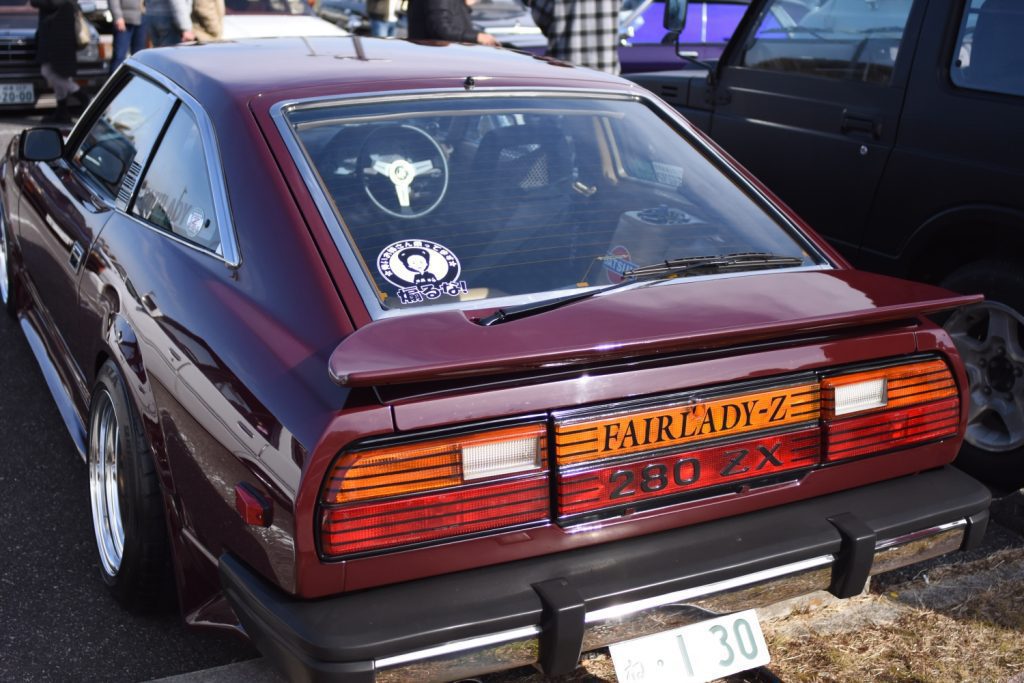 Rear view of a classic Nissan Fairlady-Z 280ZX at a car show, with a sticker reading Don't return empty-handed! in Japanese.