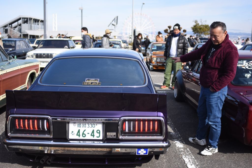 Man pointing at a Nissan Laurel car at a car show in Japan, with people and other classic cars in the background.