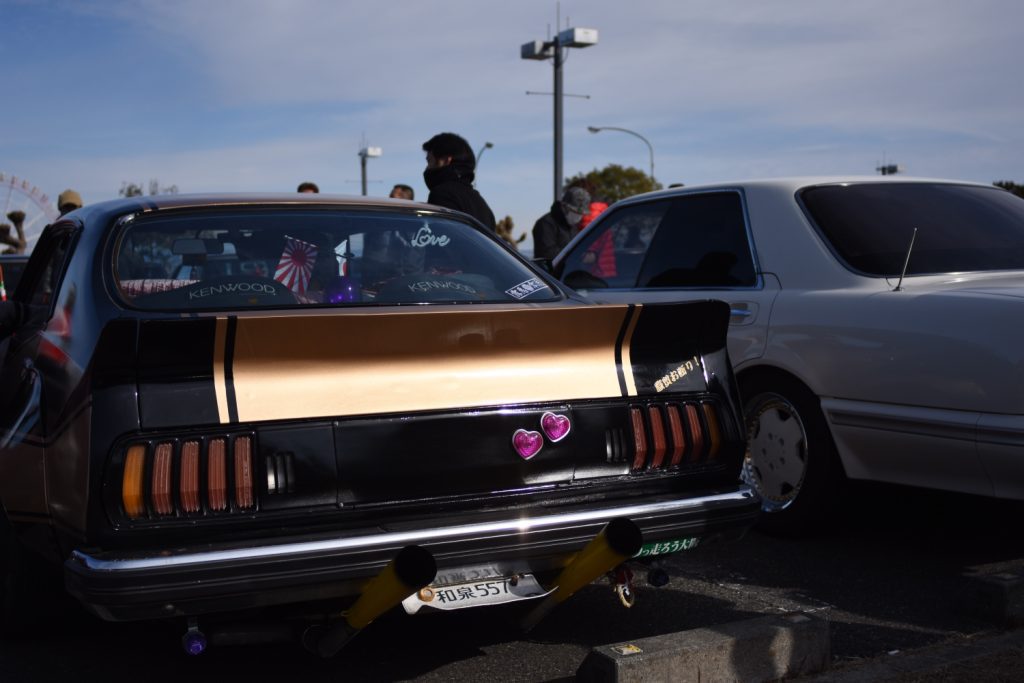 Vintage black and gold car with heart decals and Kenwood sticker at a car meet, japanese license plate visible.