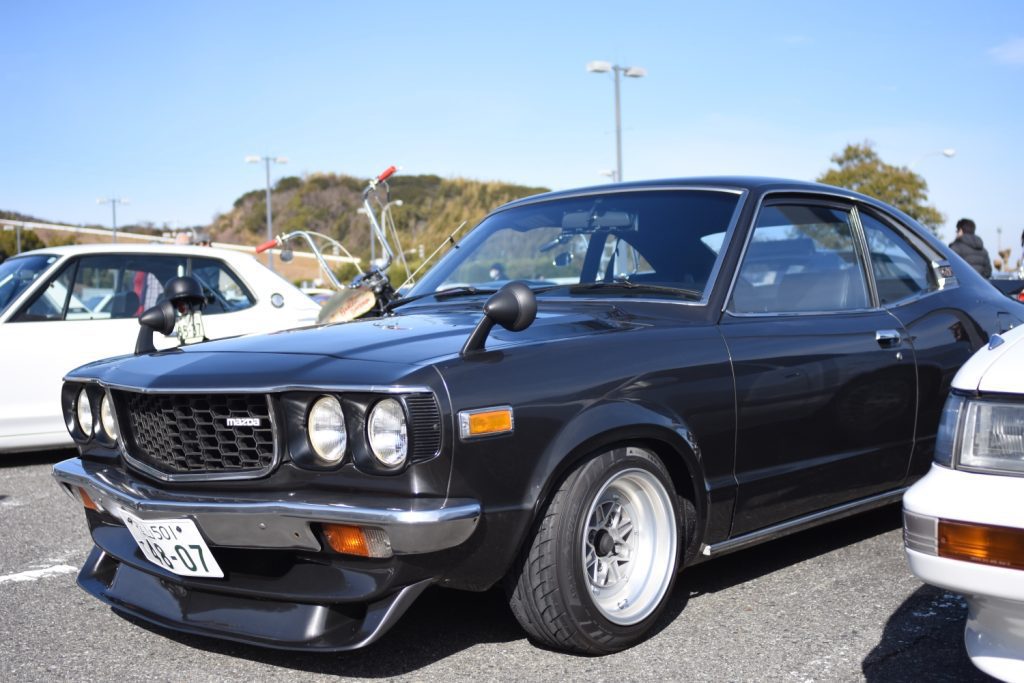 Vintage black Mazda car on display in a parking lot during a car event on a sunny day, with other vehicles in background.