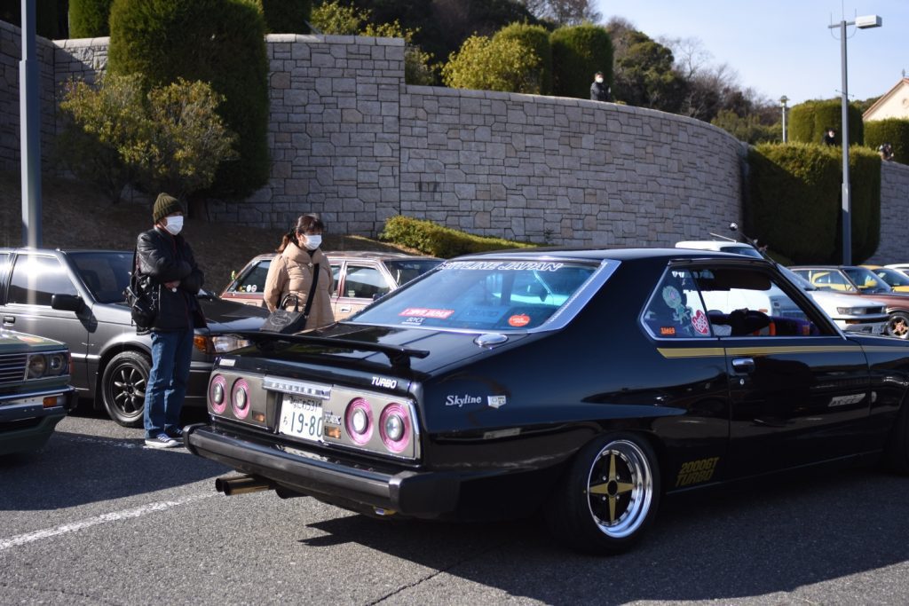 Classic black Nissan Skyline 2000GT Turbo at a car meet with spectators. License plate reads 19-80. Skyline Japan on rear window.