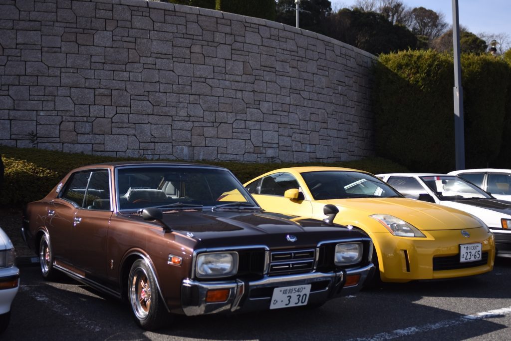 Vintage and modern Japanese cars parked beside a stone wall featuring a brown classic sedan and a yellow sports car.