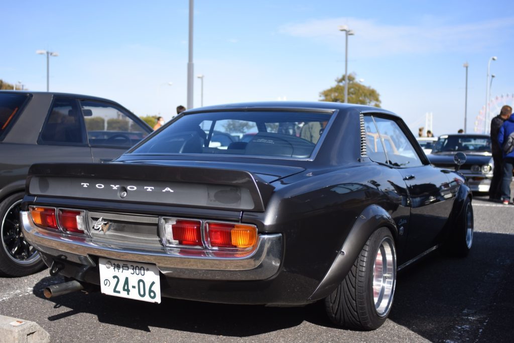 Rear view of a classic black Toyota car with Japanese license plate number 24-06 at an outdoor car show.