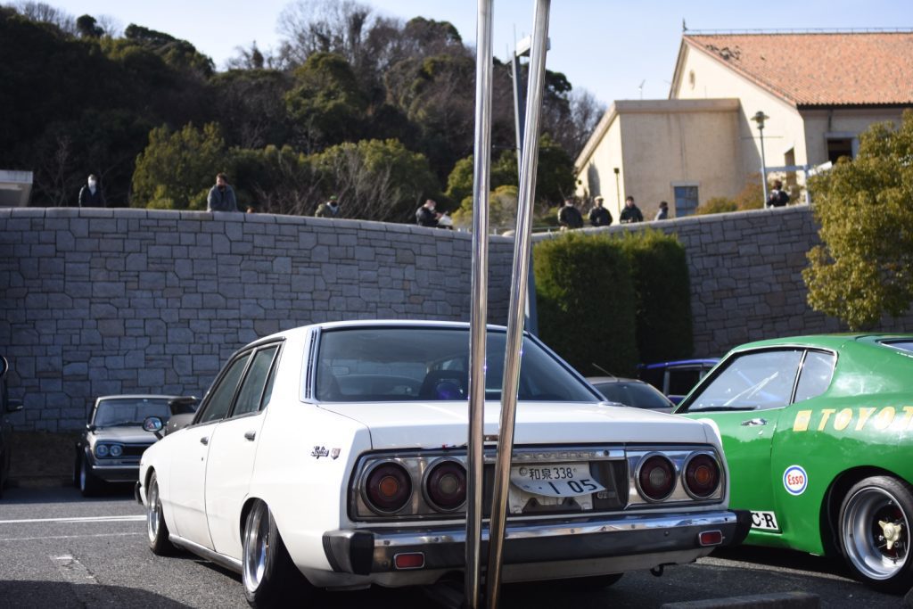 Vintage Japanese cars parked in a lot; the white one’s license plate reads 和泉 338, and the other one is a green Toyota.