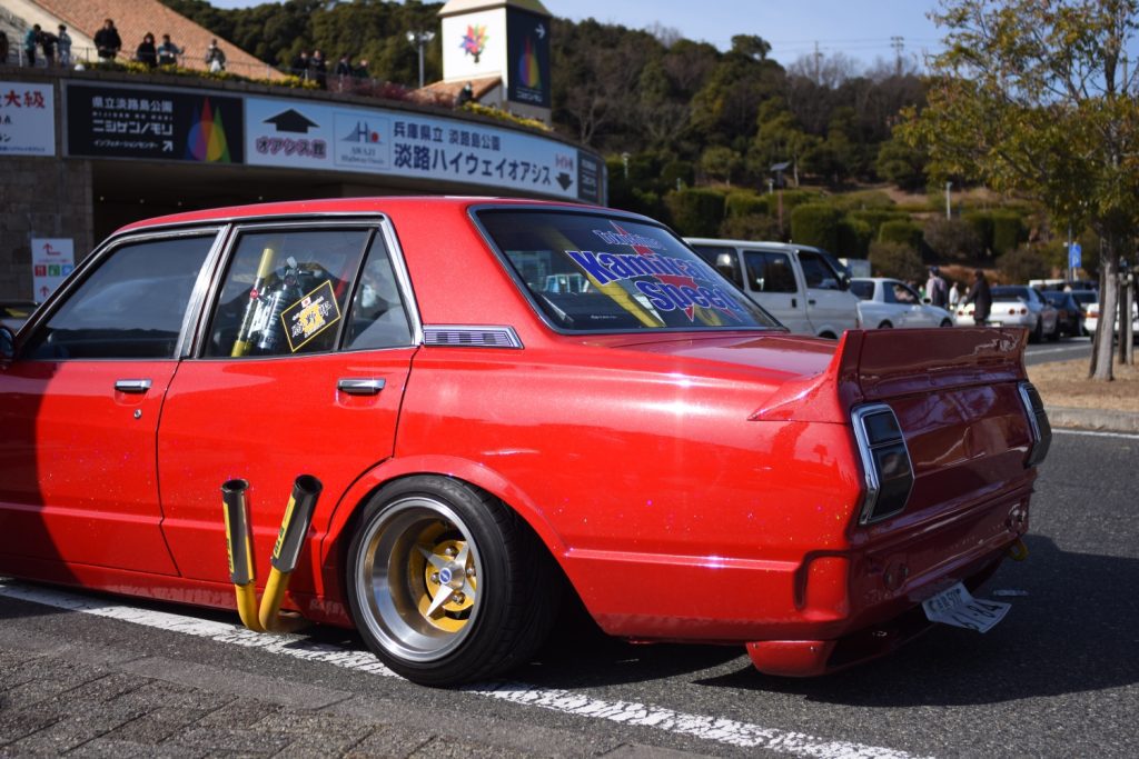 Red customized car with yellow pipes, gold rims, Kaminari Special rear window decal, and Japanese signage in the background.