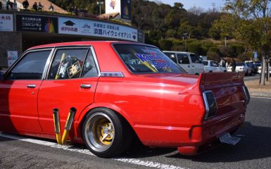 Red customized car with yellow pipes, gold rims, Kaminari Special rear window decal, and Japanese signage in the background.