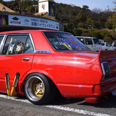 Red customized car with yellow pipes, gold rims, Kaminari Special rear window decal, and Japanese signage in the background.