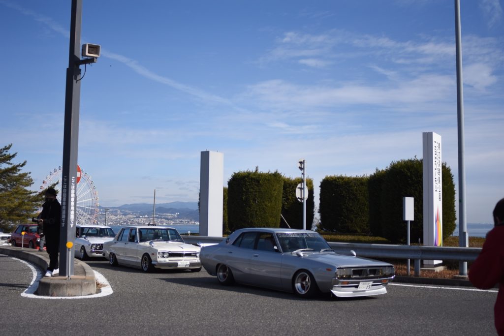 Vintage cars driving past a Ferris wheel at Biwako Terrace in Shiga, Japan, on a clear day with blue skies.