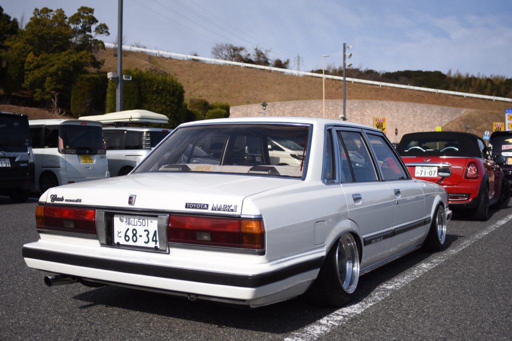 Classic white Toyota Mark II in a parking lot, low stance, custom wheels, with Japanese license plate reading 68-34.