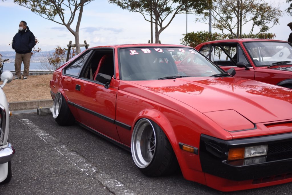 A red lowered sports car with wide wheels at a car meet. Japanese text on windshield: Showa Work.