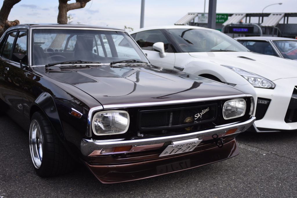 Classic Nissan Skyline parked next to modern sports cars at a car event, showcasing iconic Japanese automotive design.