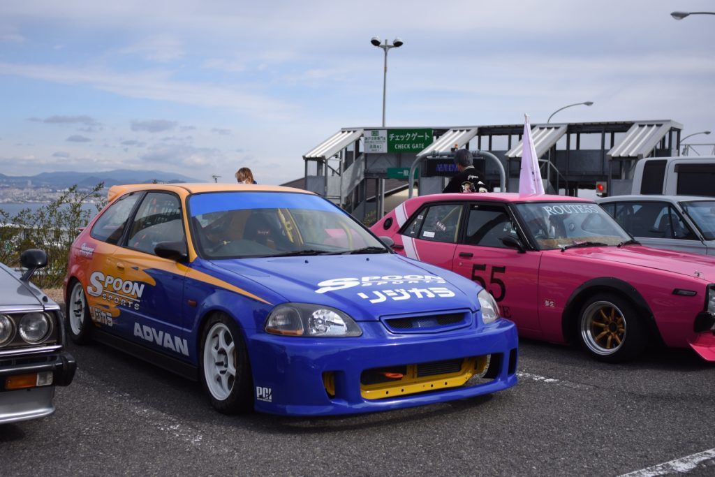 Modified blue and yellow Honda Civic with Spoon Sports logo parked next to a pink car at a car meet.