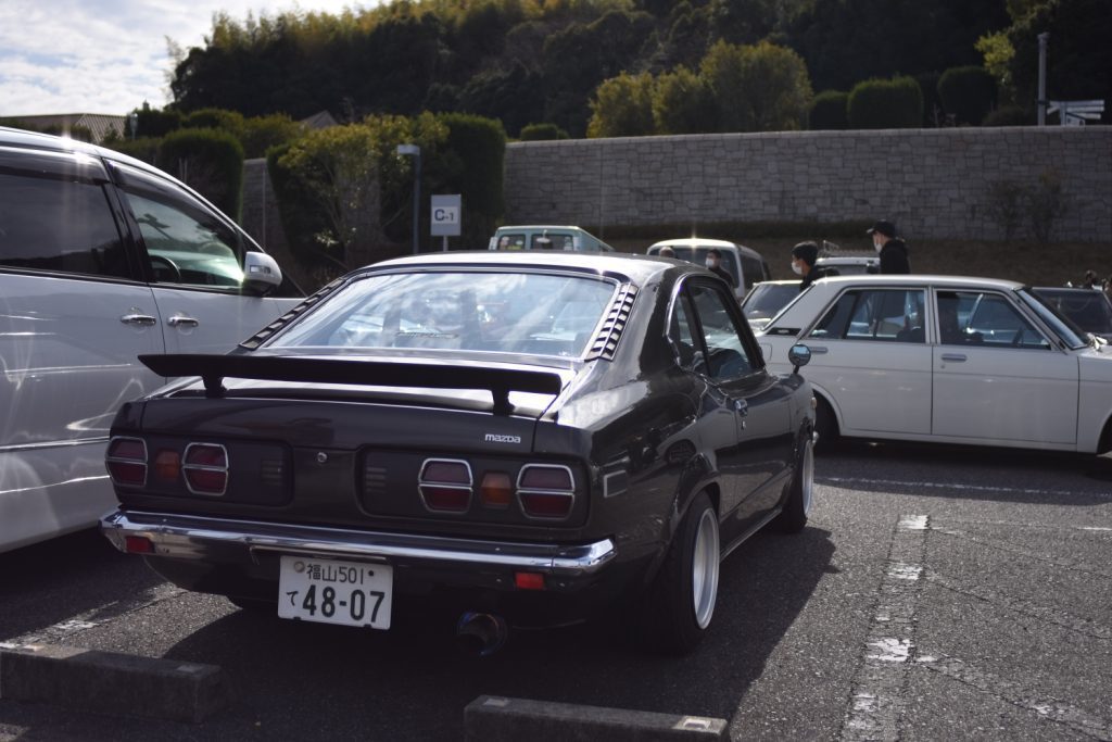 Classic black Mazda with Fukuyama license plate in parking lot, alongside other vintage cars under a sunny sky.