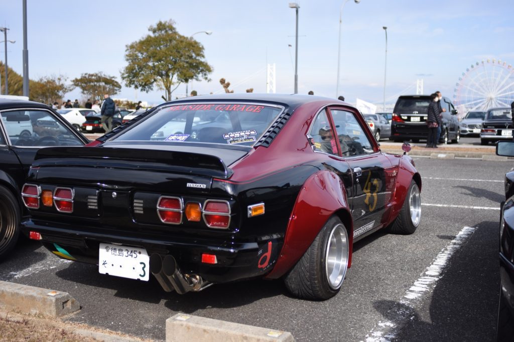 Vintage modified Mazda RX-3 with Seaside Garage decal, parked at classic car event in Japan, showcasing its sporty design.