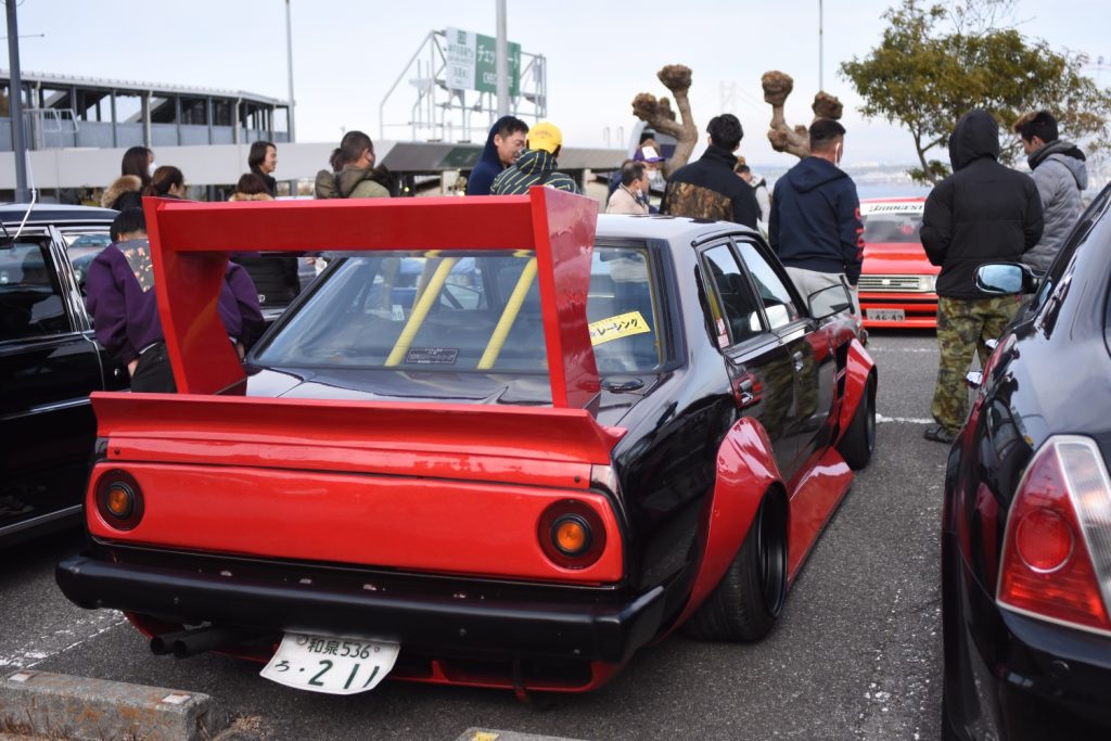 Custom black and red car with large rear wing at car meet in Japan. Sign in window reads Racing.