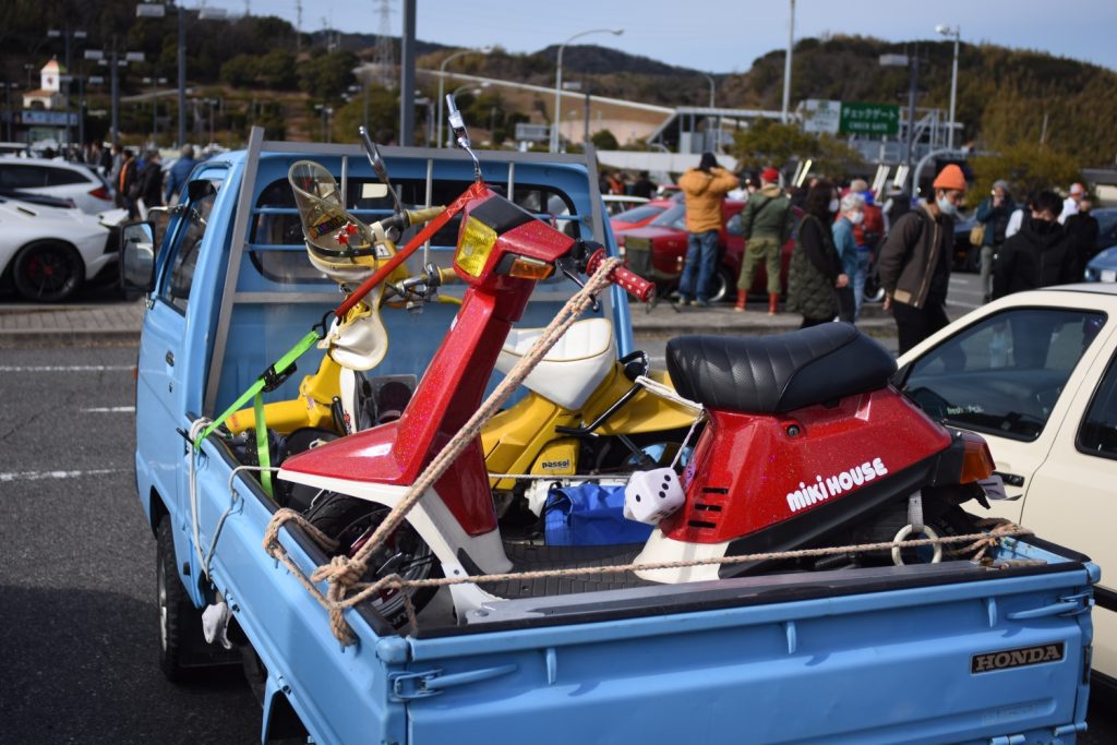 A blue truck carrying two scooters, a red Miki House and a yellow Passol, at a crowded parking lot.