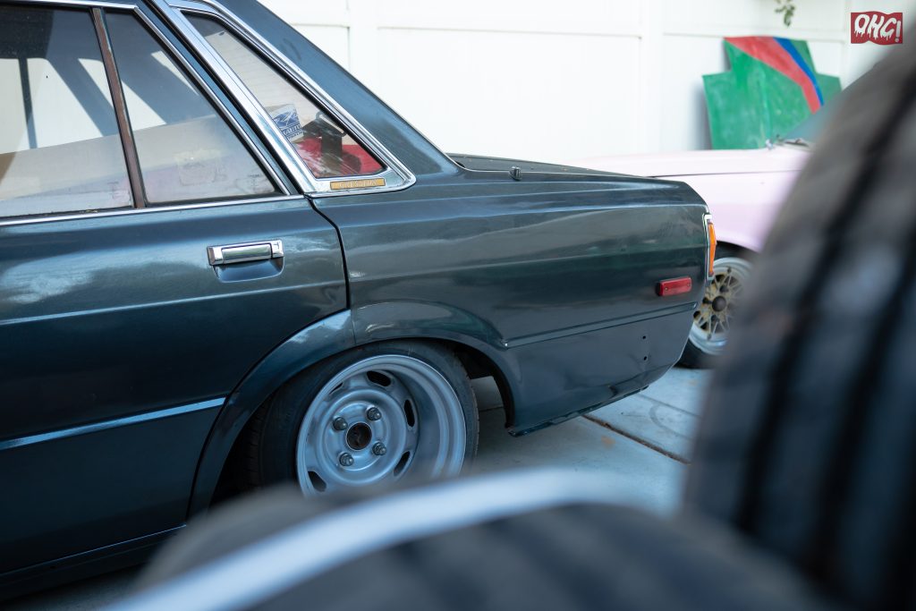Close-up of a classic dark green sedan with the badge CRESSIDA on the rear quarter panel parked next to a pink vehicle.