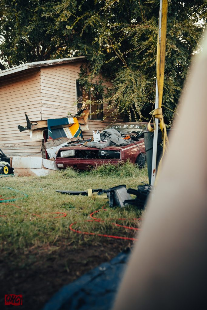 Abandoned car with Japanese text (team 初夢会), scrap materials, and overgrown plants by a wooden shed in a cluttered yard.