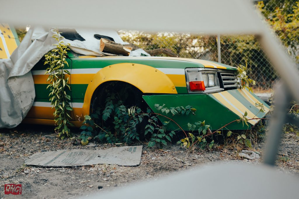Abandoned yellow and green race car overgrown with plants, partially covered by a tarp in a fenced area. オハギ (Ohagi) logo visible.