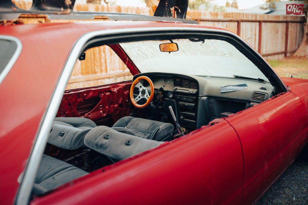 Interior of a dismantled red car with orange steering wheel, featuring Japanese sticker New Beautiful Boy on the dashboard.