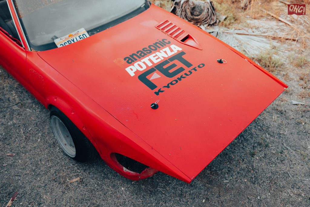 Vintage red car in California with Panasonic and Potenza branding on the hood, parked on a grassy area.