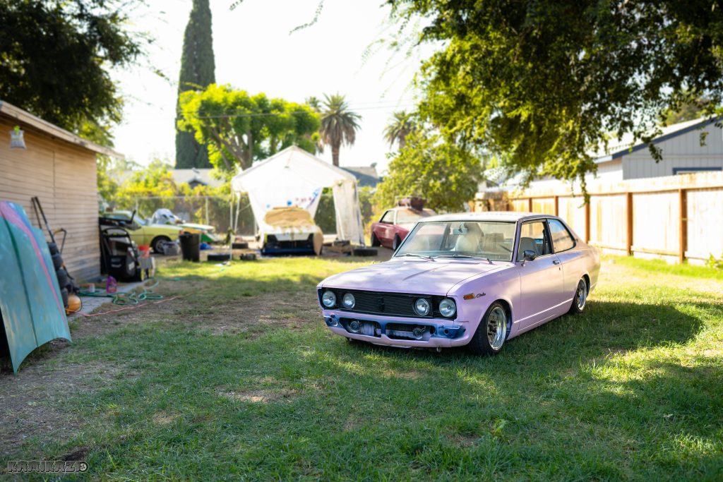 Vintage purple car parked in a sunny backyard with a canopy in the background and vibrant green surroundings.