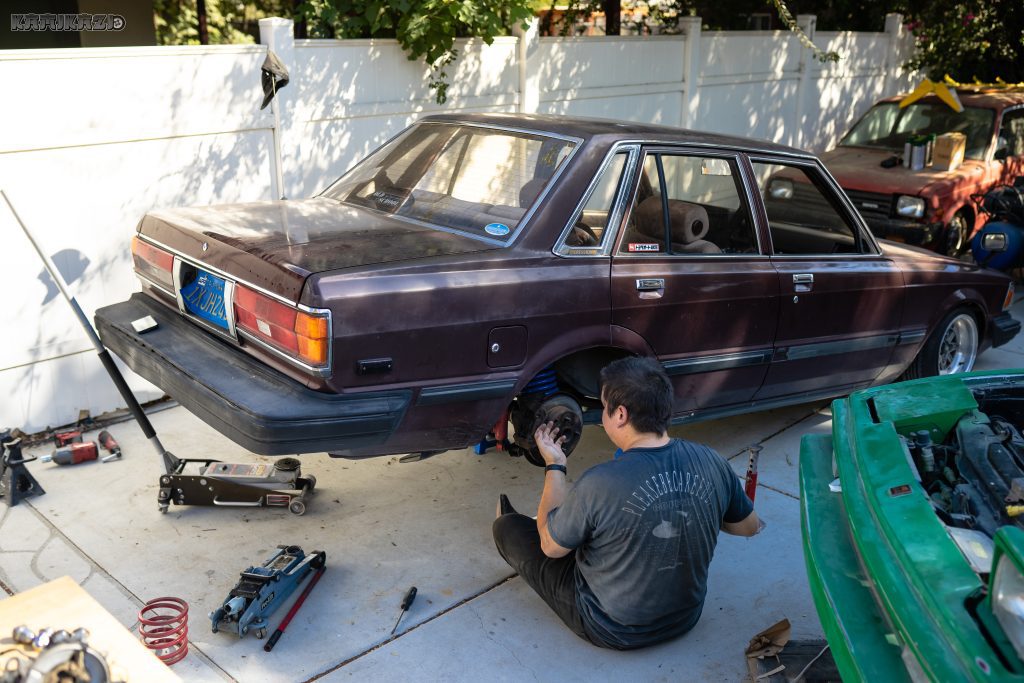 A man works on a vintage car's rear brakes in an outdoor garage with tools and equipment scattered around.