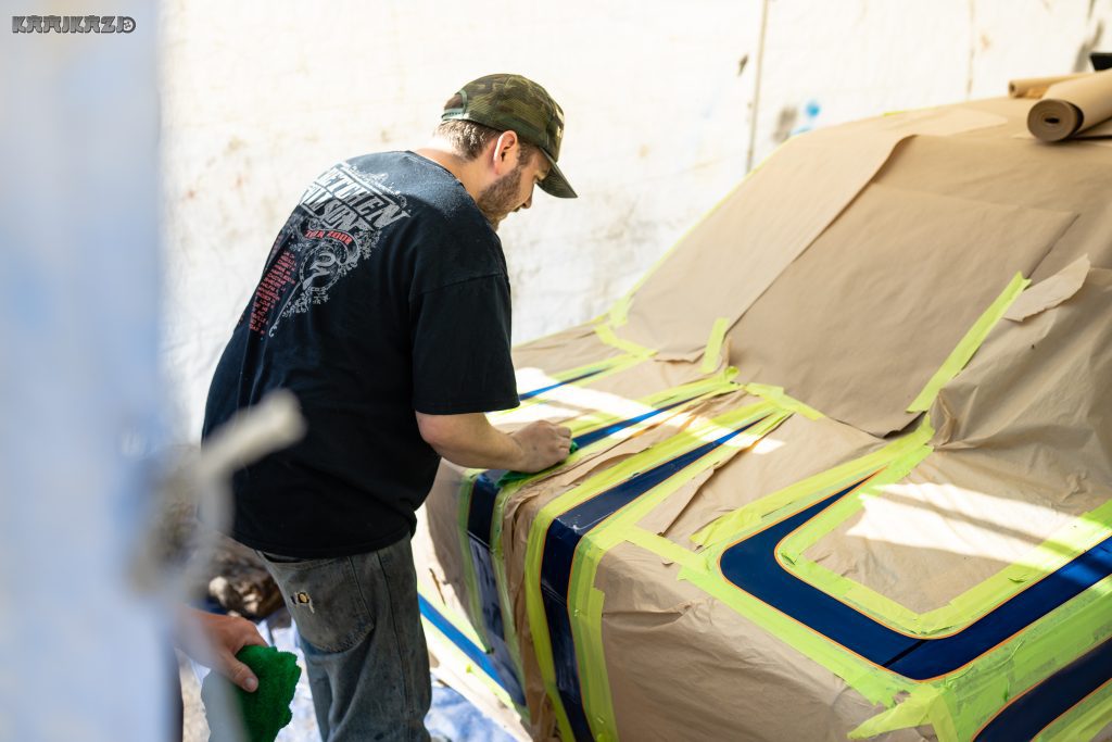 A man in a camo hat and black shirt works on a taped, paper-covered car in a workshop. Japanese word: カミカゼ (Kamikaze).