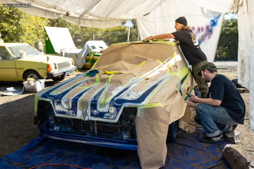 Two mechanics work on a classic car under a tarp, masking it for a new paint job. Old yellow car visible in the background.