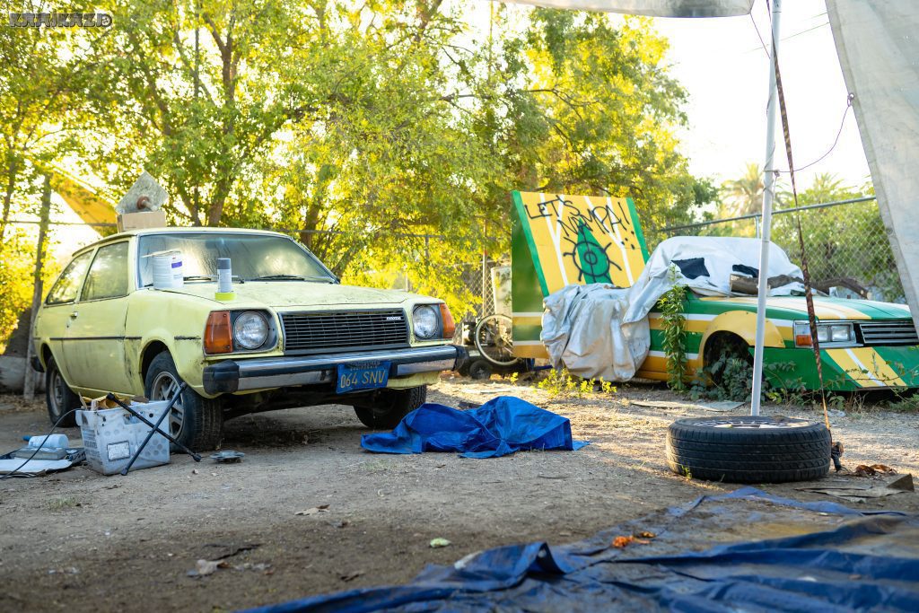 Two vintage cars in a rustic outdoor setting with a vibrant Let's Go! sign in the background.