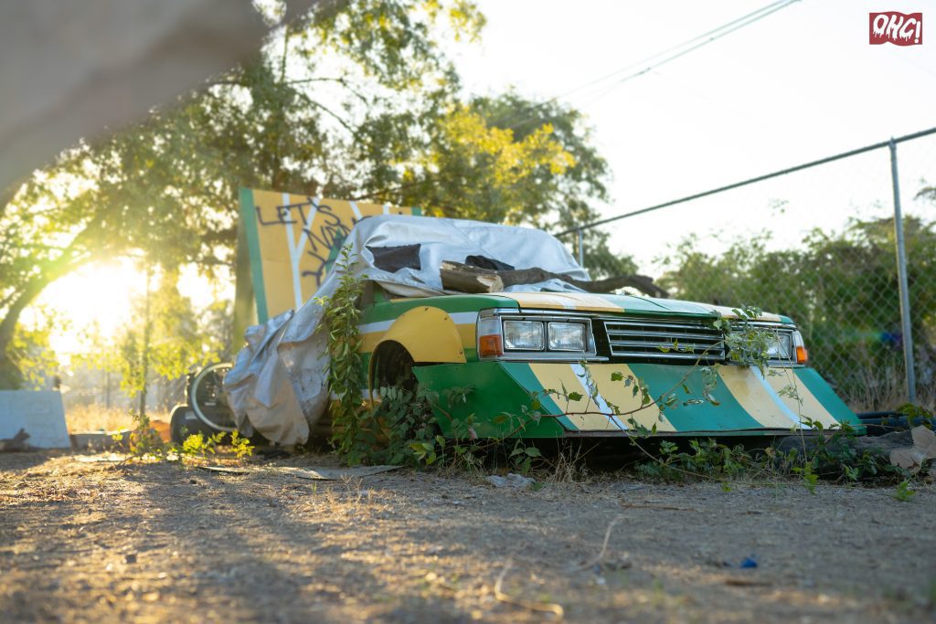 Abandoned car covered with tarp at sunset in a fenced area, overgrown with plants, graffiti in the background.